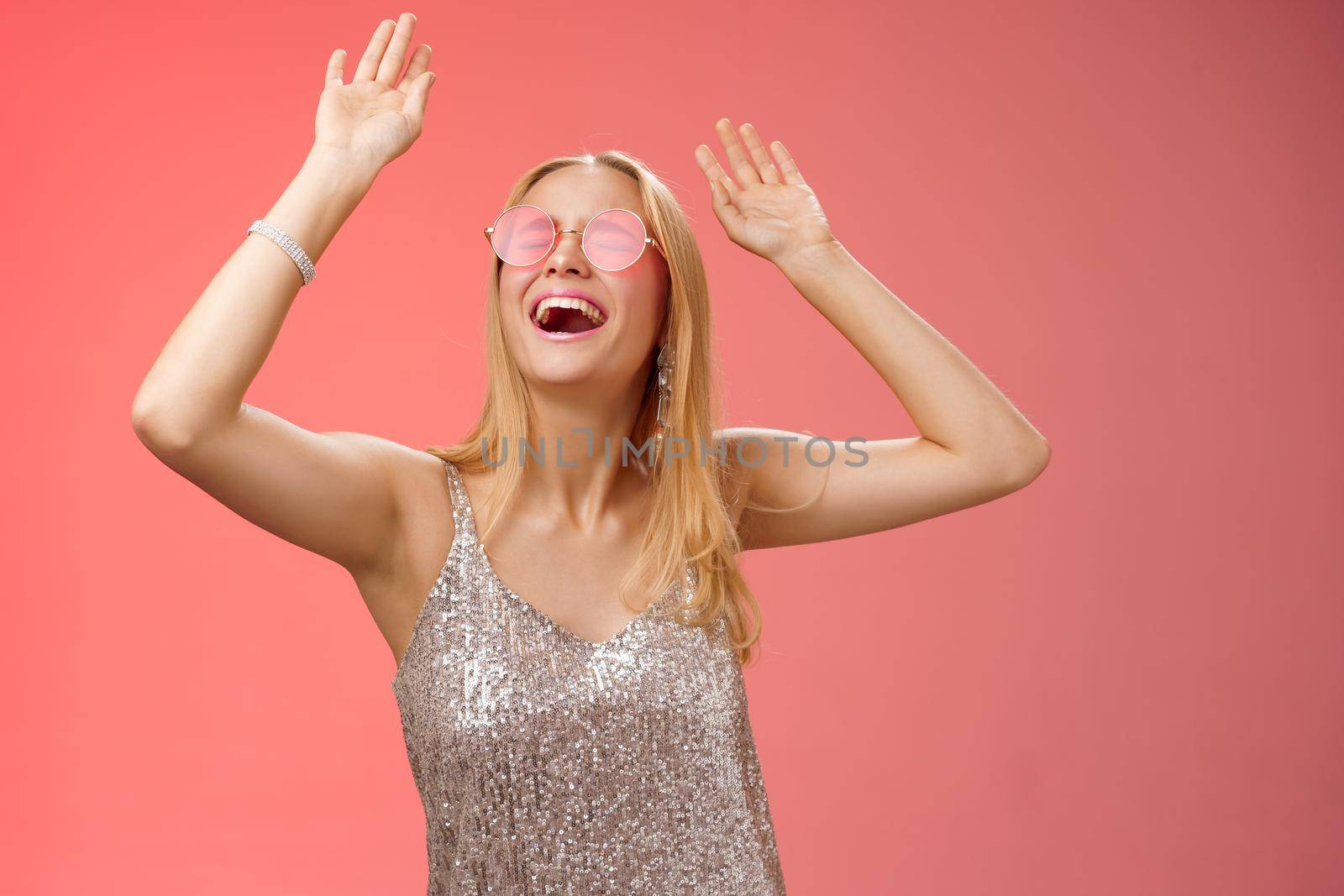 Excited chilling energized young blond woman in silver stylish glittering dress sunglasses raise hands up having fun dancing dance-floor nightclub throw party celebrate b-day, red background.