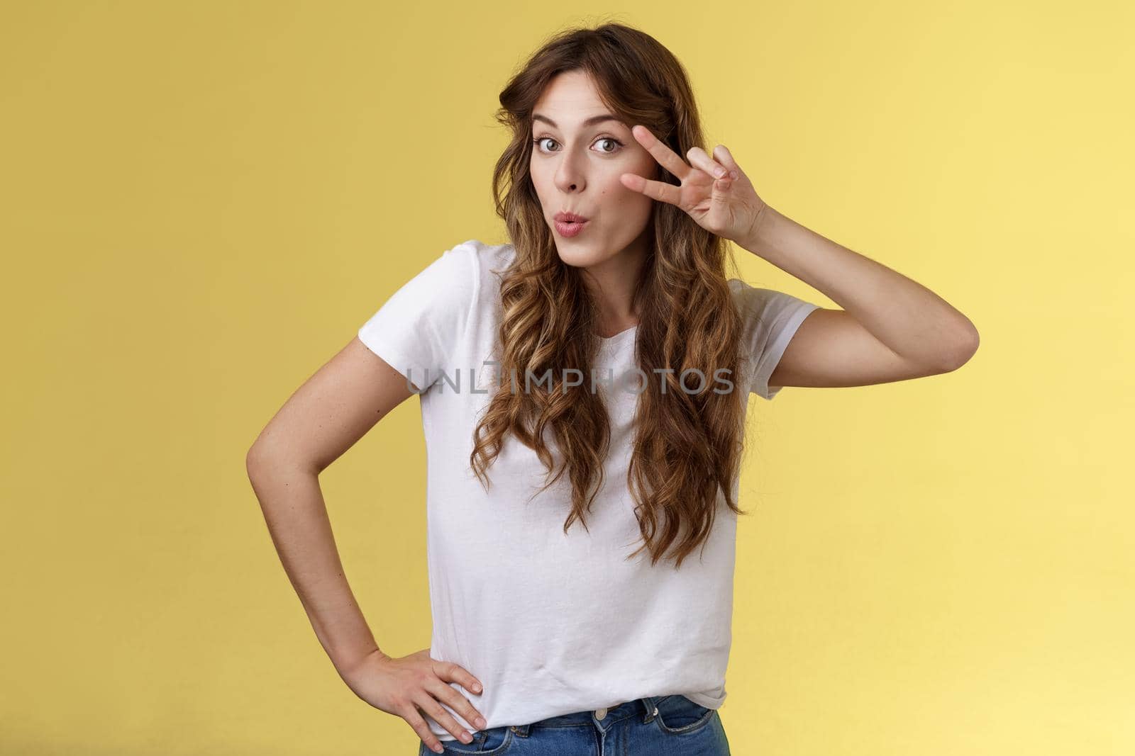 Cute glamour young woman curly haircut tilt head silly show peace victory sign fold lips lovely muah kiss gesture send flirty glances camera hold hip mimicking sensually gazing you yellow background. Lifestyle.