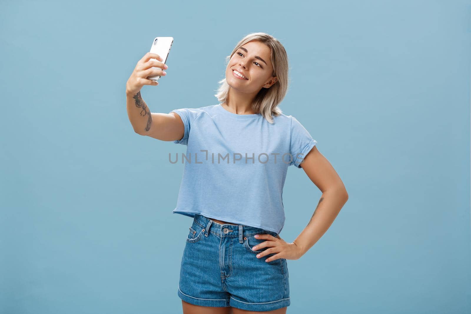 Stylish cute female gym manager taking selfie with new smartphone posing with broad pleased smile and holding hand on hip making post for followers standing carefree and relaxed over blue wall by Benzoix