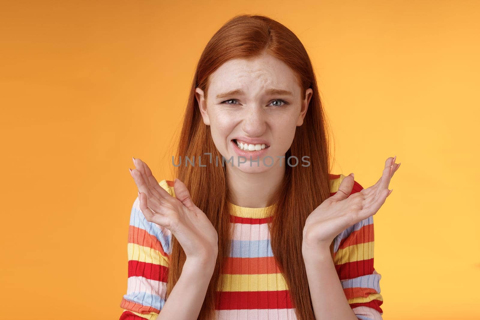 Awkward unhappy worried young redhead girl cringe feel sorry apologizing smirking smiling nervously frowning squinting spread hands sideways shrugging confused, standing orange background.