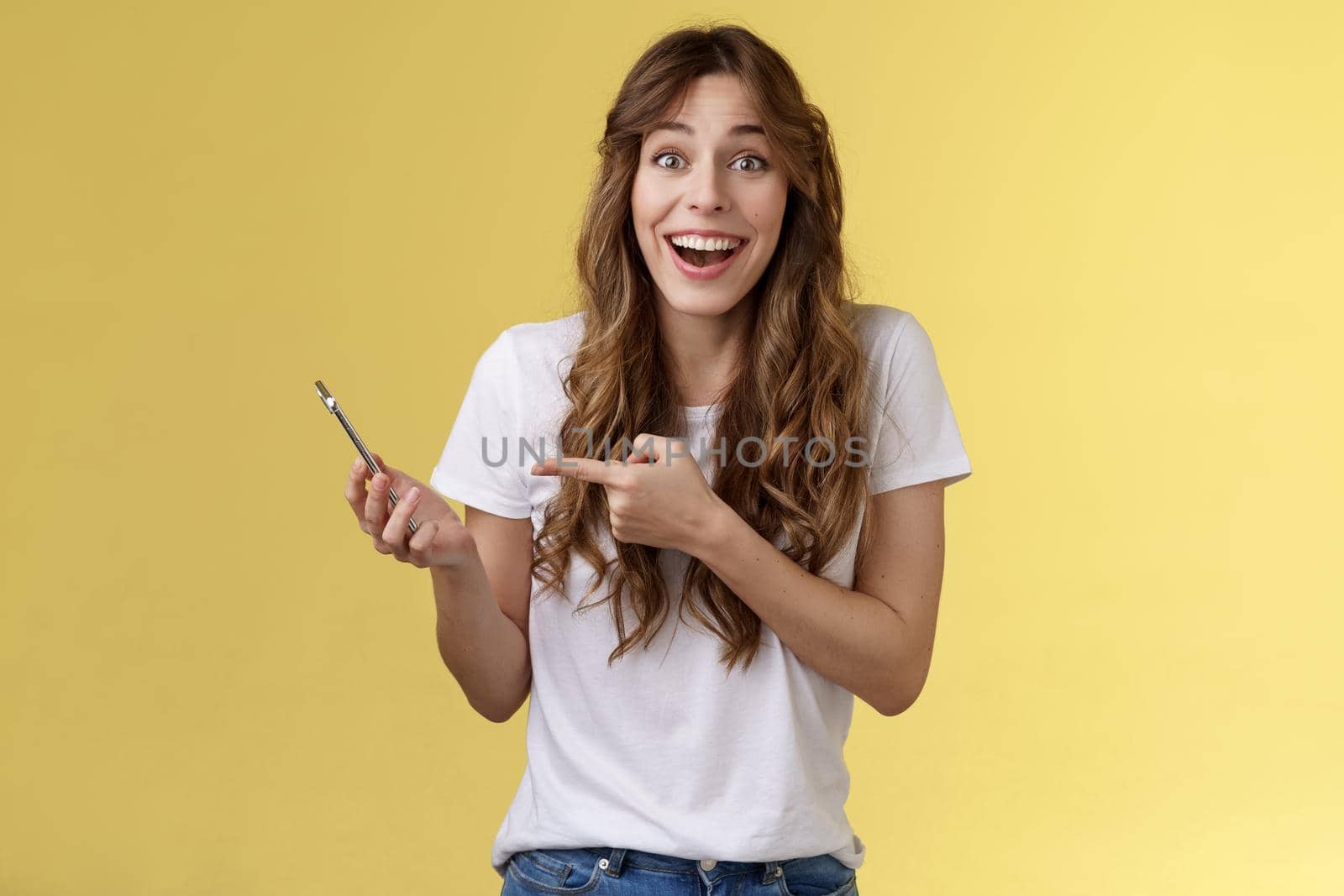 Cheerful happy attractive caucasian female curly hairstyle smiling laughing surprised receive pleasant good news via phone call holding smartphone describe awesome post read internet pointing mobile by Benzoix