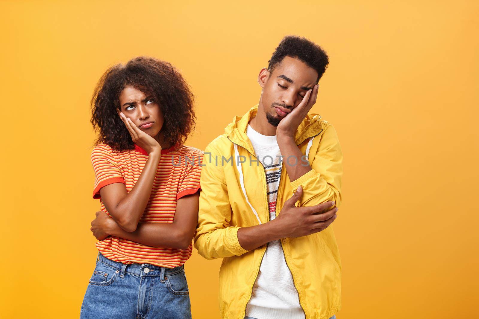 Girl feeling displeased and offended on boyfriend who fell asleep during date pursing lips frowning looking up while boyfriend leaning head on face with closed eyes and tired look over orange wall by Benzoix