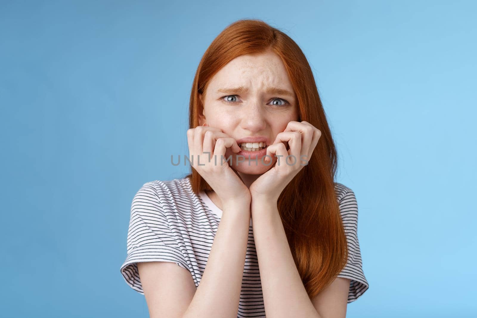 Worried uncomfortable scared young panicking redhead young girl feeling pressure distressed frowning squinting frightened biting fingernails trembling fear, standing blue background by Benzoix