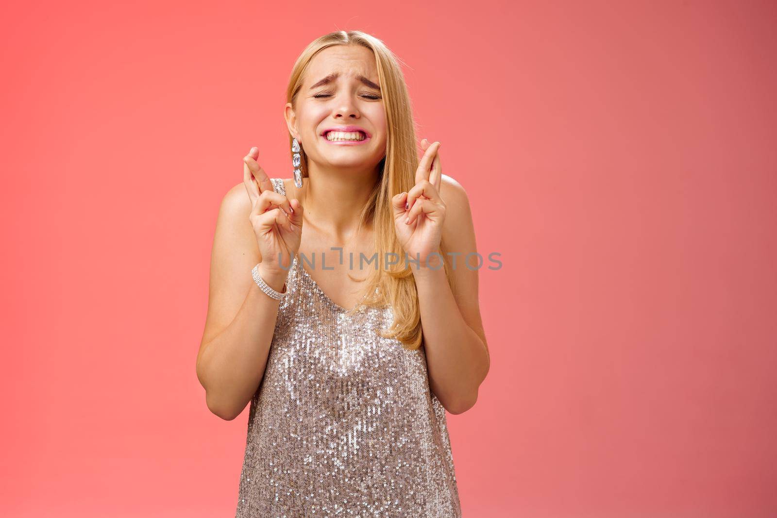 Nervous hopeful blond young woman in silver trendy dress close eyes grimacing eager wish come true cross fingers good luck praying supplicating anticiapting fortune, standing worried red background.