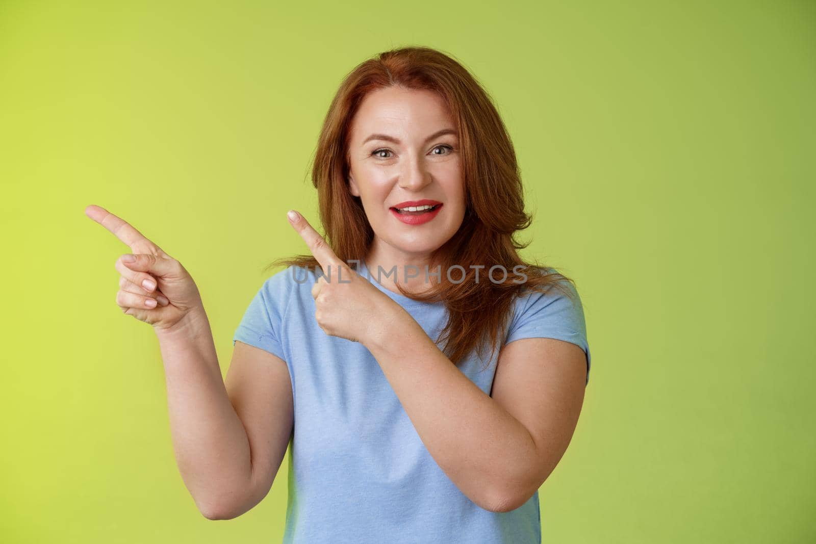 Curious enthusiastic gorgeous redhead female intrigued pointing upper left corner asking question interesting product talk shop assistant consulting promo stand green background blue t-shirt.