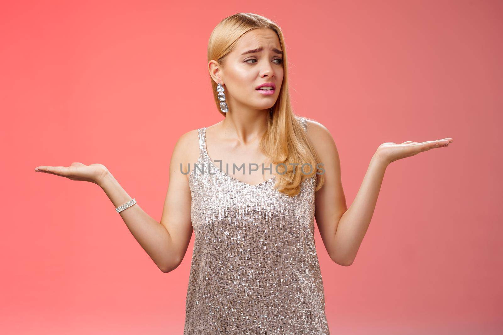Anxious insecure young cute blond girl feel lost unsure shrugging hands sideways weighing decisions look right seek help standing lost nervous hesitating red background in silver glittering dress by Benzoix