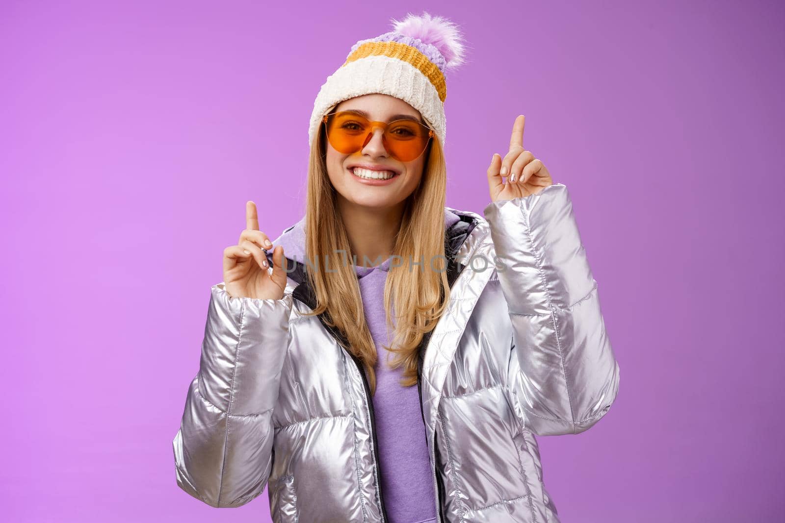 Joyful energized entertained cute blond woman having fun enjoy vacation snowy mountain trip wearing sunglasses silver jacket winter hat dancing pointing up amused standing purple background by Benzoix