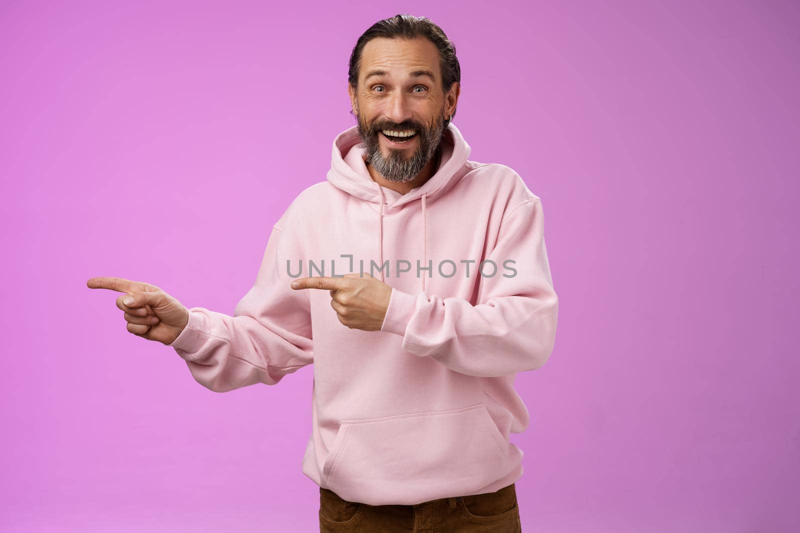 Charismatic happy good-looking mature 50s man wearing stylish hipster hoodie smiling excited pointing left index fingers impressed awesome interesting proposal standing pleased purple background.