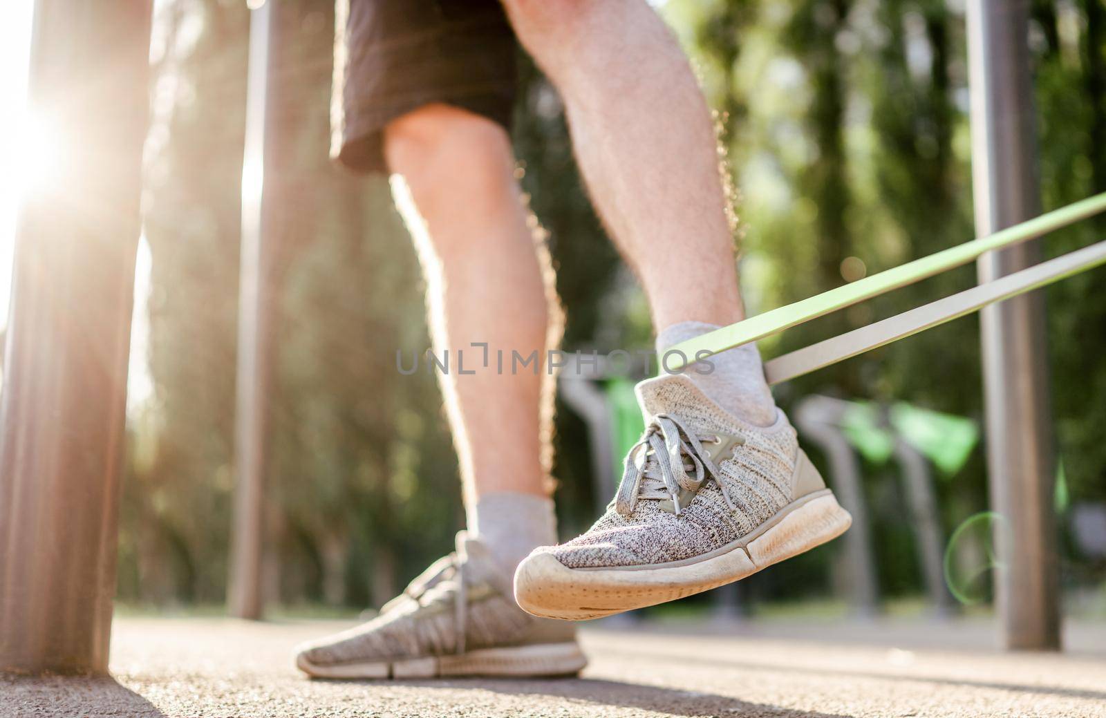Closeup side view of legs feets of man guy training with elastic rubber band at the stadium outdoors. Adult male person during workout with additional sport equipmant in summertime