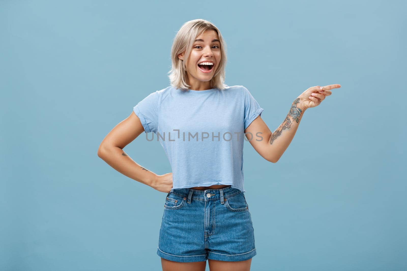 Studio shot of impressed and surprised entertained blonde female in summer outfit holding hand on waist pointing right and smiling joyfully being amazed and amused over blue background. Emotions and advertisement concept