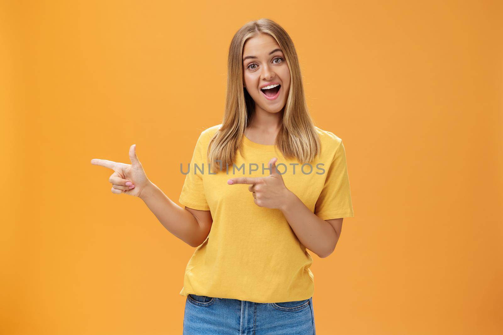 Check out copy sapce of dream. Upbeat good-looking pleasant female shop assistant in t-shirt pointing left with finger guns and smiling amused at camera recommending to watch that direction by Benzoix