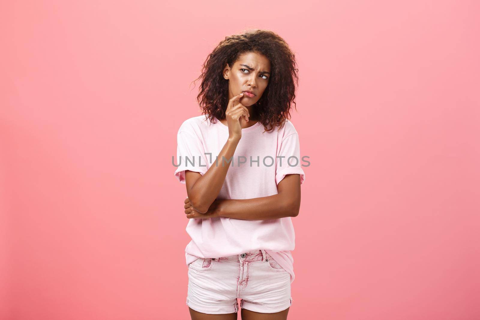 Suspicious displeased dark-skinned young curly-haired woman in trendy t-shirt and shorts saying hmm touching chin with finger frowning and staring left doubtful and unsure having concern. Emotions concept