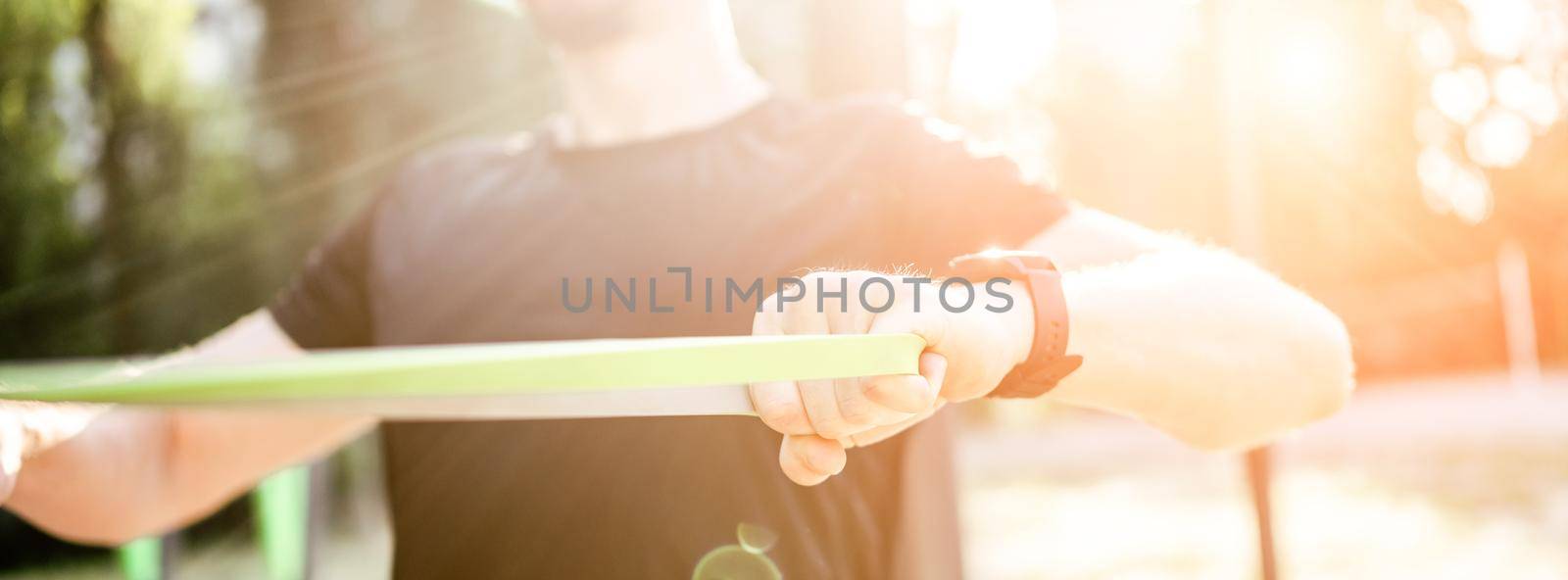 Strong man guy training with elastic rubber band at the stadium outdoors. Adult male person during workout for arms and chest with additional sport equipmant. Sportsman exercising outside