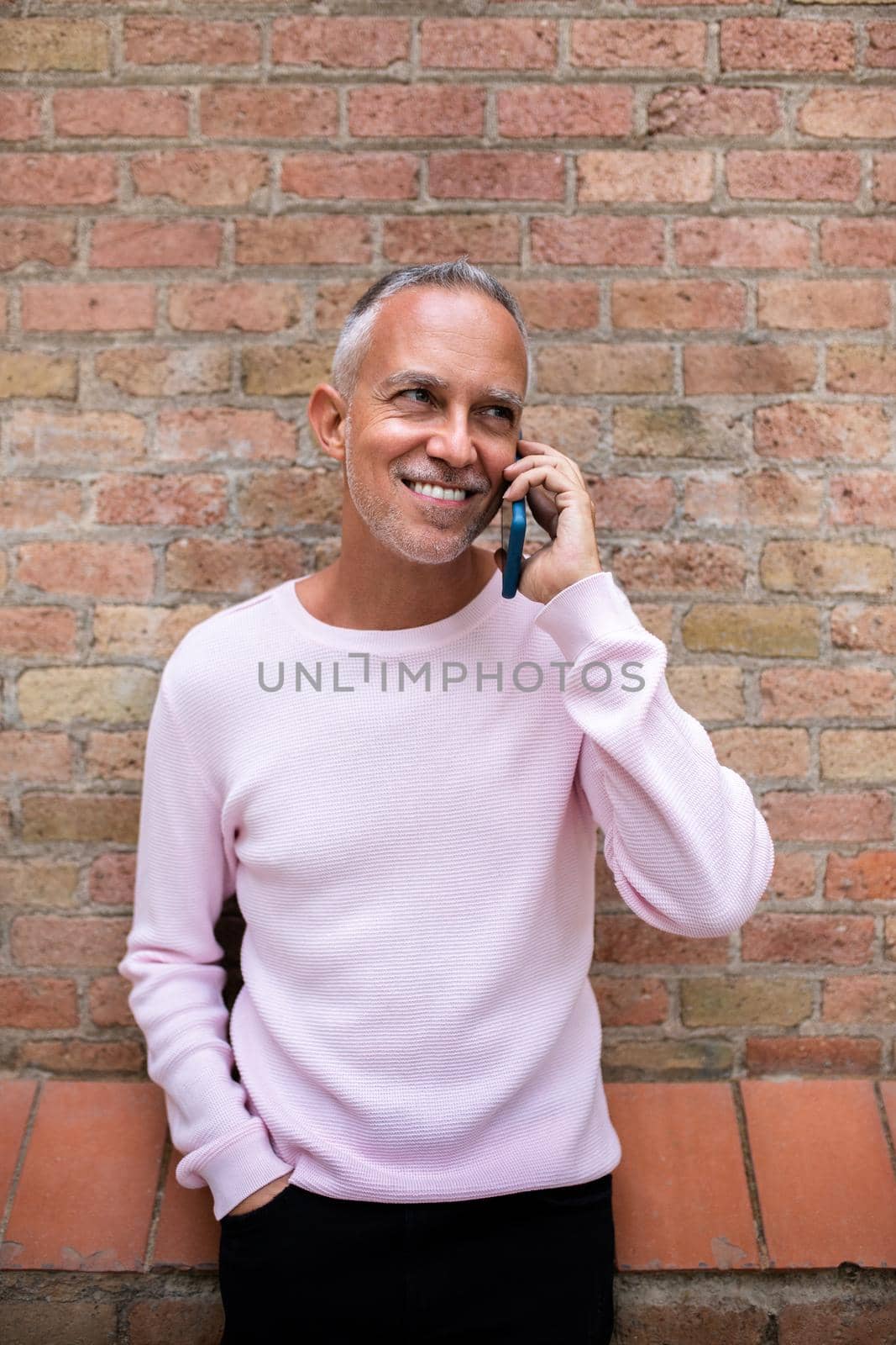 Smiling caucasian man talking with mobile phone leaning against orange brick wall. Vertical image. Lifestyle and technology concept.