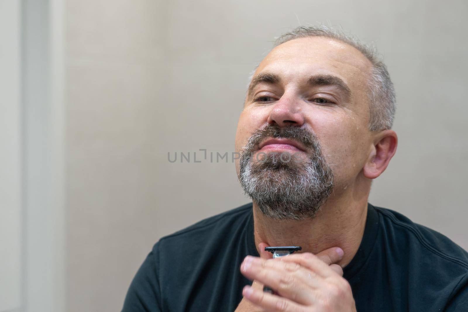 Charming middle aged man shaving his beard and moustache off in front of mirror to look younger. Mature gray-haired man restyling his beard himself at home using razor