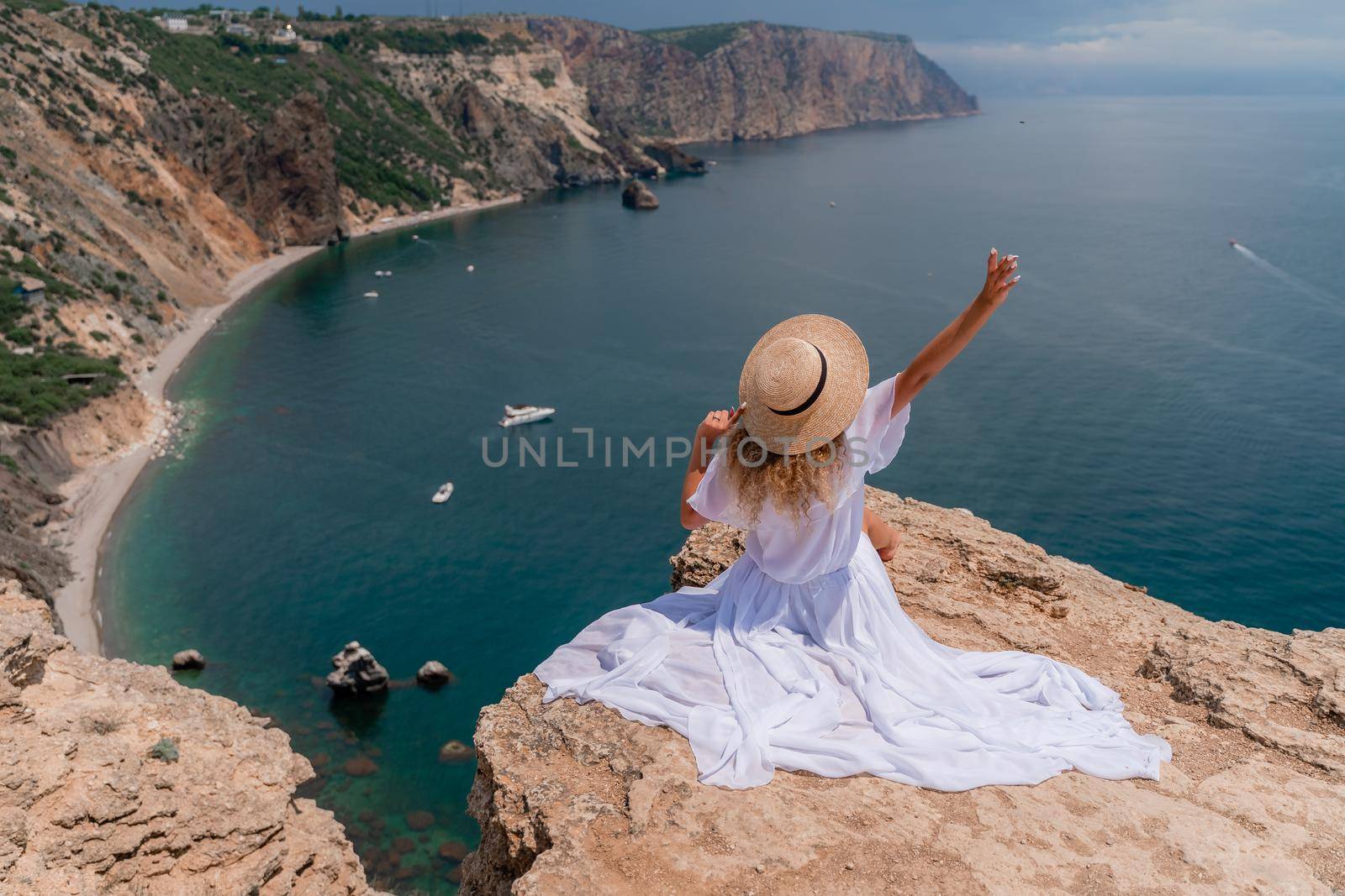 Portrait of a young woman on the beach by the sea, sitting with raised hands in a white dress and hat. Back photo.