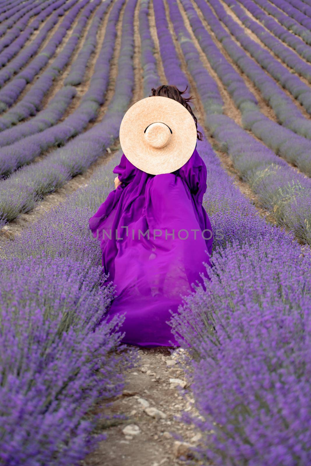 A young beautiful girl in a purple flying dress stands on a blooming lavender field. Rear view. The model has a straw hat by Matiunina