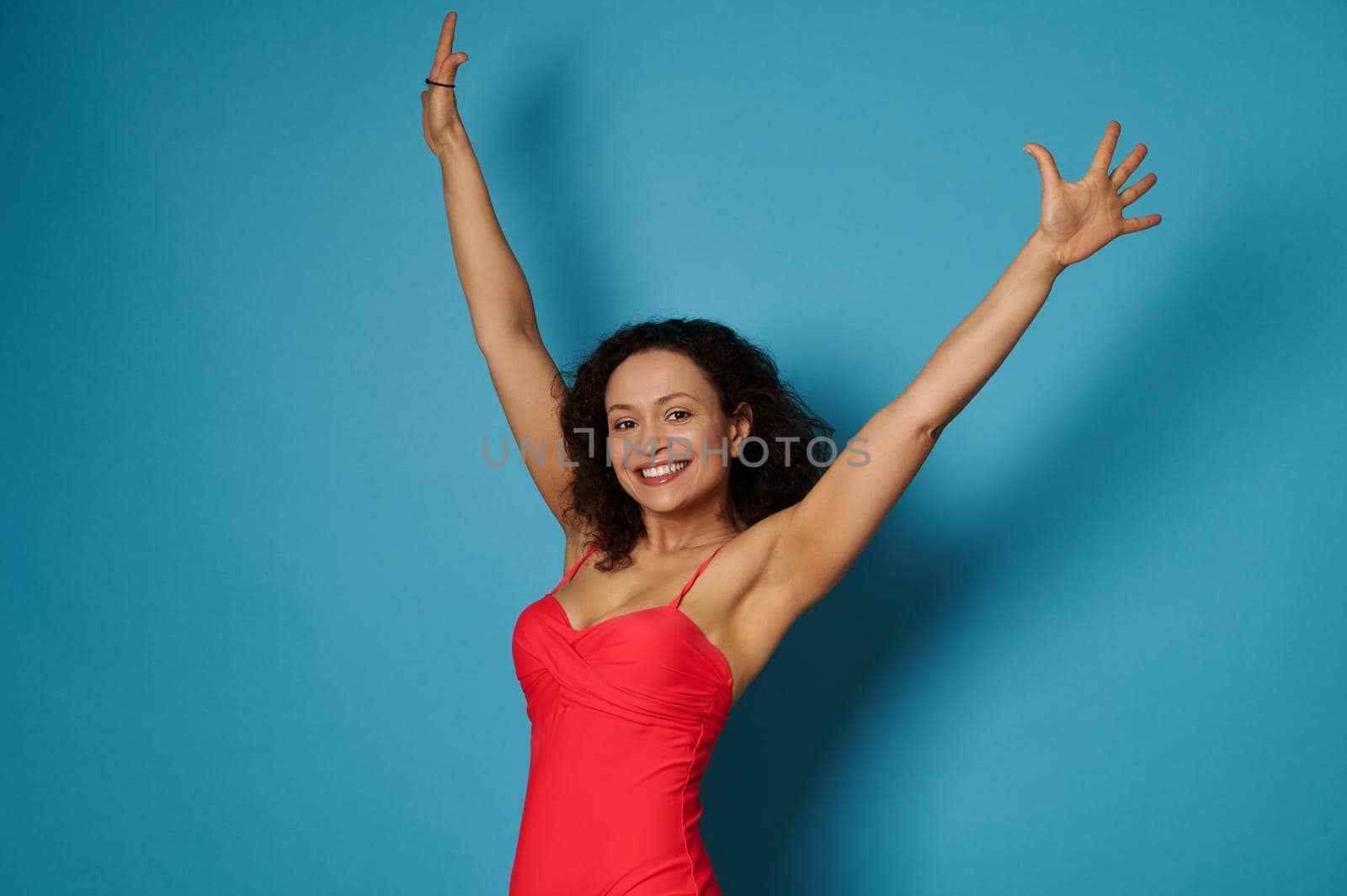 Smiling African American woman in red swimsuit posing with arms raised on blue background with copy space