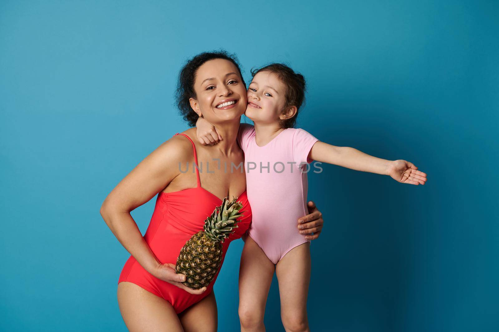 A cute little girl hugging her mother with a pineapple in her arms, both in bathing suits. Happy mothers day and summer concepts on blue background.