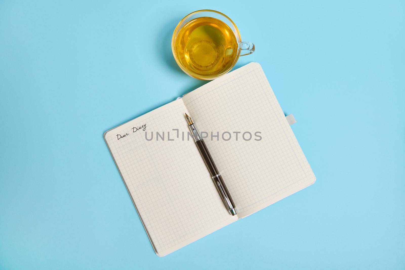 Open notepad with word Dear Diary and an ink pen next to a tea cup on blue background with copy space. High angle view, flat lay.