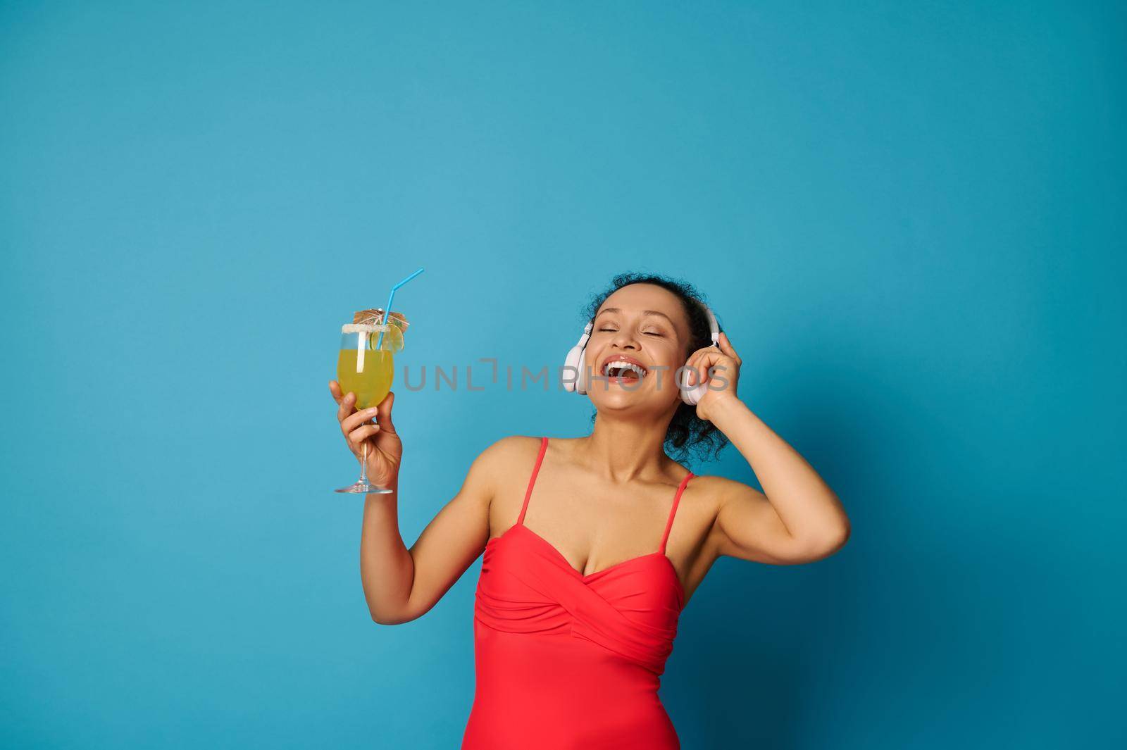 Happy brunette with closed eyes holding a summer cocktail and enjoying listening to music with headphones by artgf