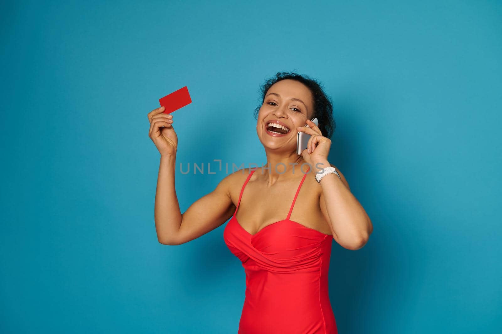 Cute brunette with beautiful smile talking on smartphone and holding a red blank card while smiling to the camera, isolated on blue background with copy space