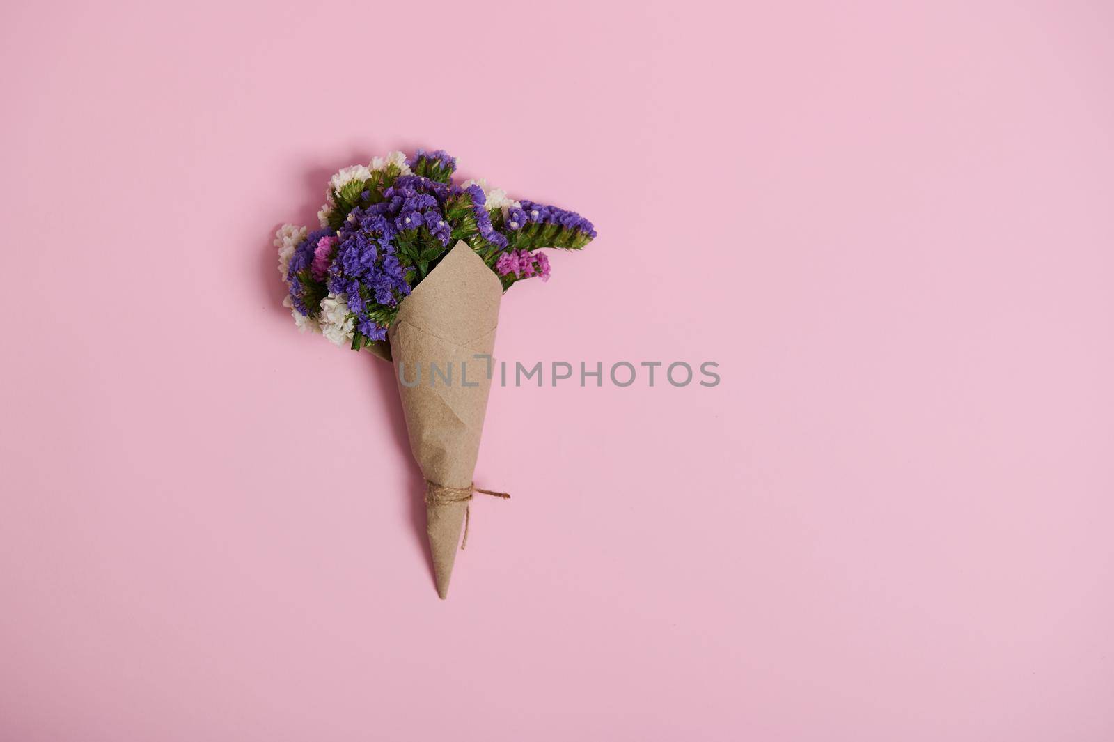 Flat lay of a floral composition of beautiful meadow flowers wrapped in wrapping craft paper tied with rope, isolated on pink background with copy space.