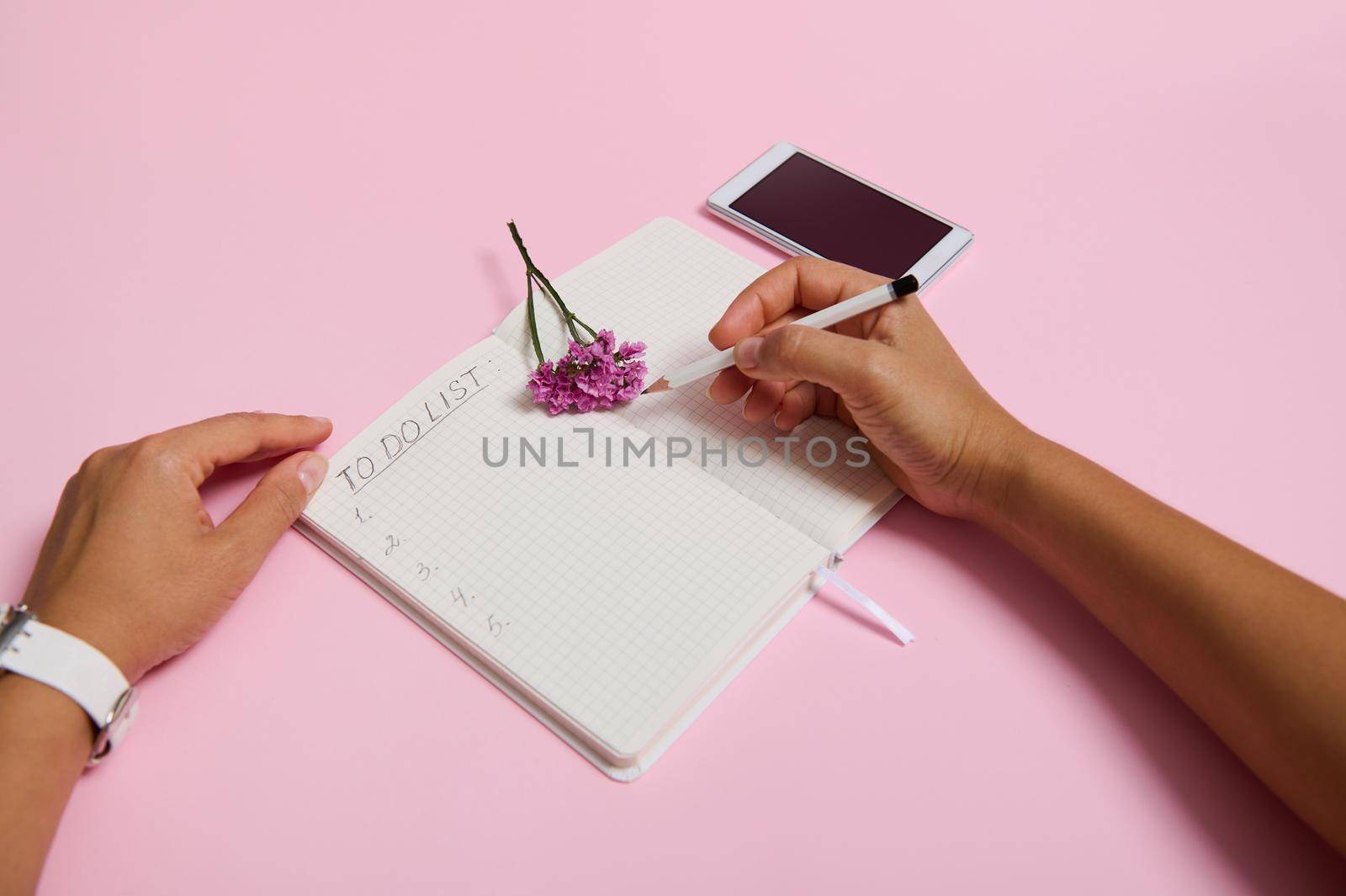 Top view of female hands holding pencil, writing in notebook, checking to-do list. Mobile phone and pink flower lying on pink background with copy space. Business, planning and time management concept