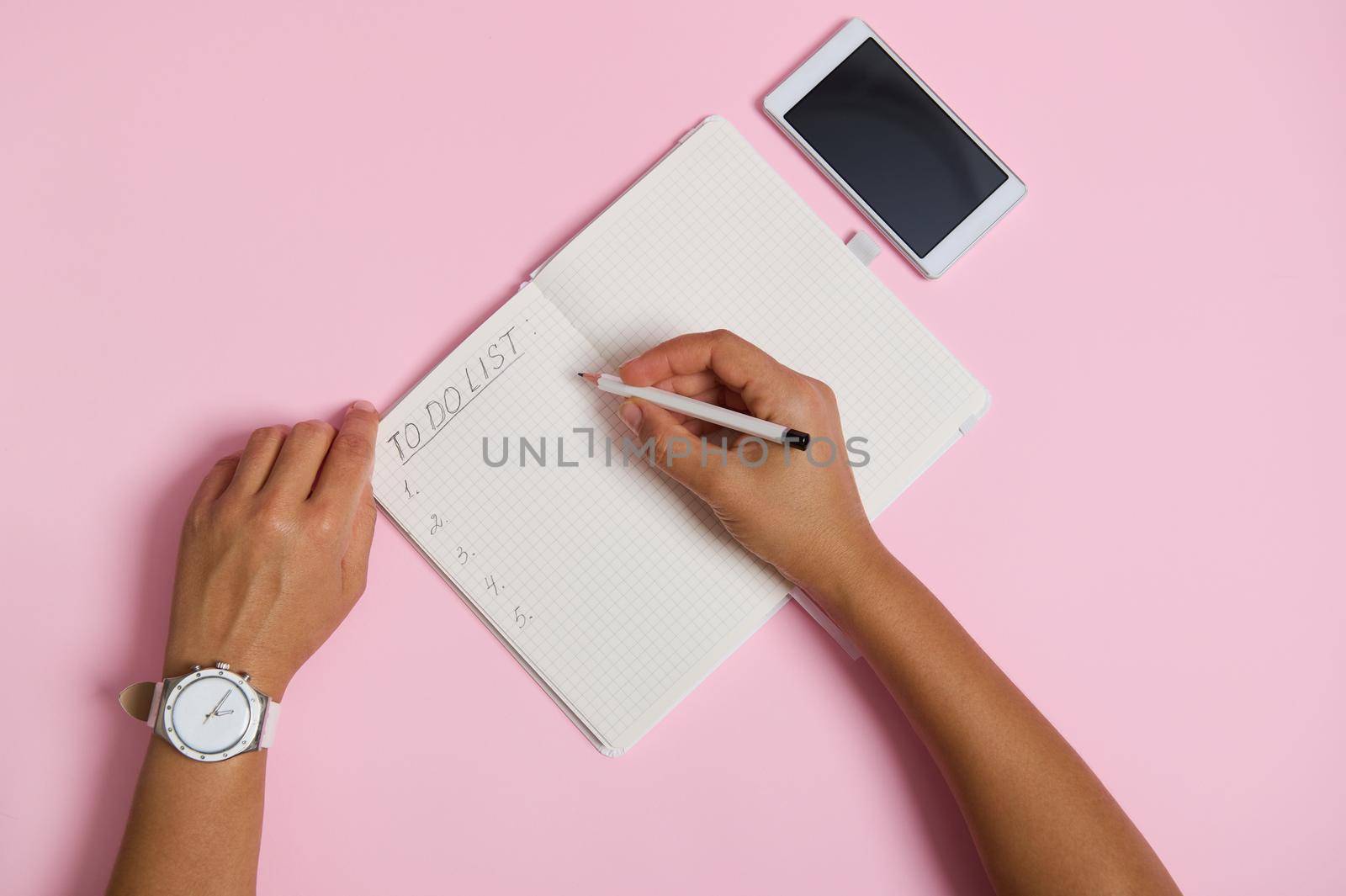 High angle view of female hands holding pencil, writing in notebook, checking to-do list. Mobile phone lying on pink background with space for text by artgf