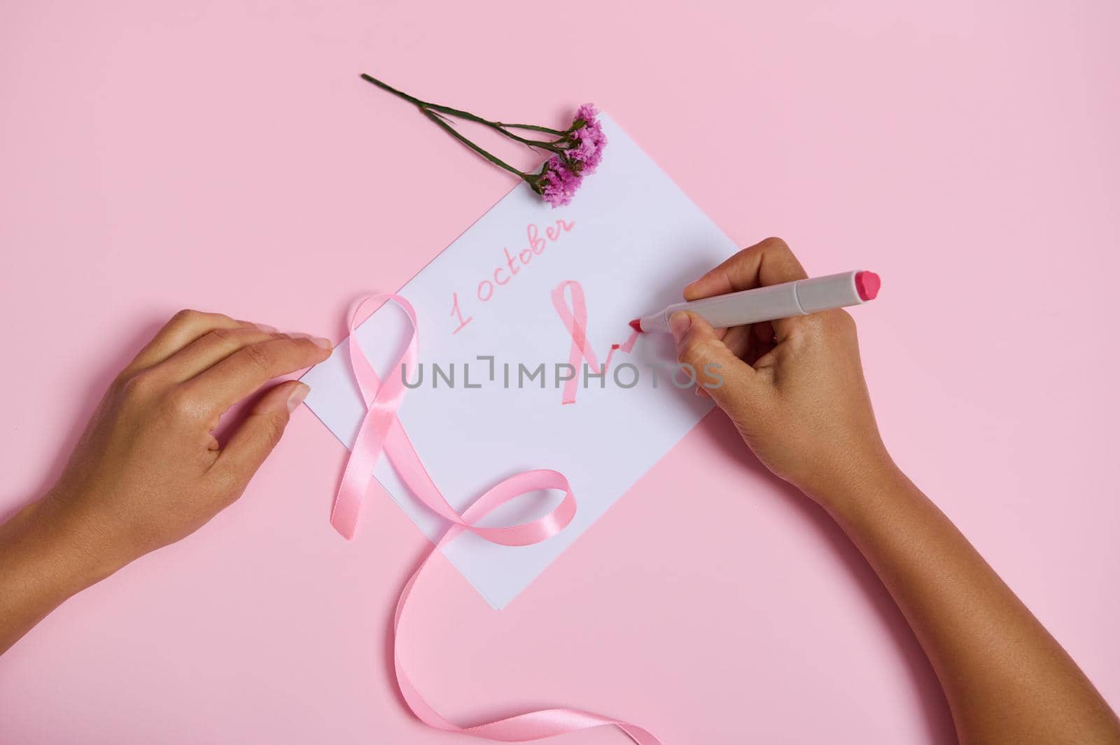 Woman's hand holds a felt-tip pen writes october 1 and draws on the paper a pink symbol of Breast Cancer Awareness Month, a pink ribbon with an endless end, lying on a pink background