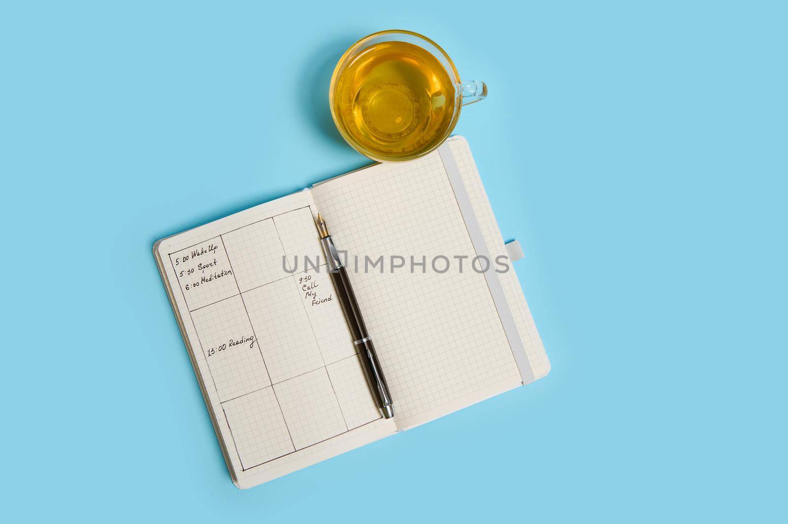 Flat lay composition of an open agenda with schedule, ink pen and transparent glass cup of tea lie on blue surface. Color background with copy space . Time management, deadline, schedule concept