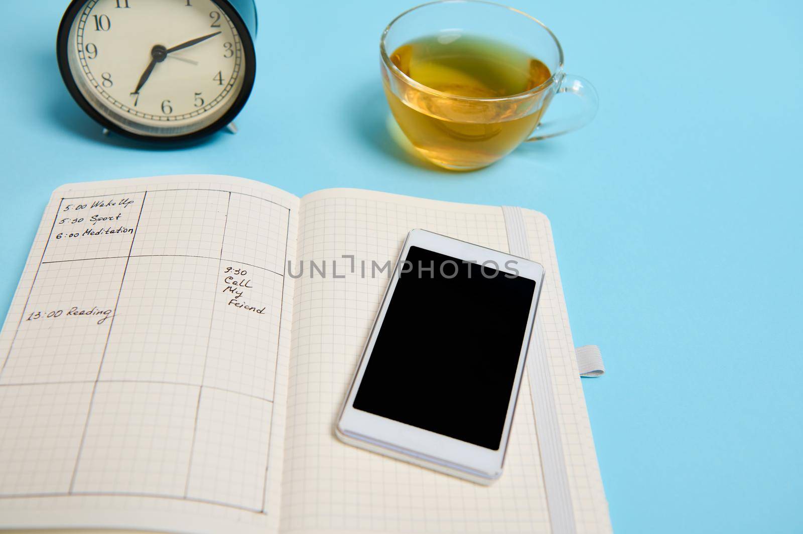 An open notebook with schedule, smartphone, glass cup of tea and black alarm clock lie on blue surface. Color background with copy space. Time management, deadline and schedule concept