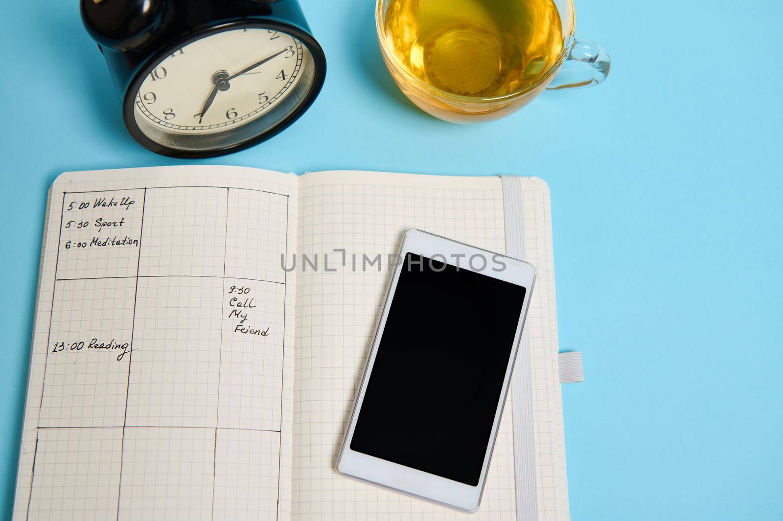 Flat lay of open notebook with schedule, smartphone, glass cup of tea and black alarm clock lie on blue surface. Color background with copy space . Time management, deadline, schedule concept
