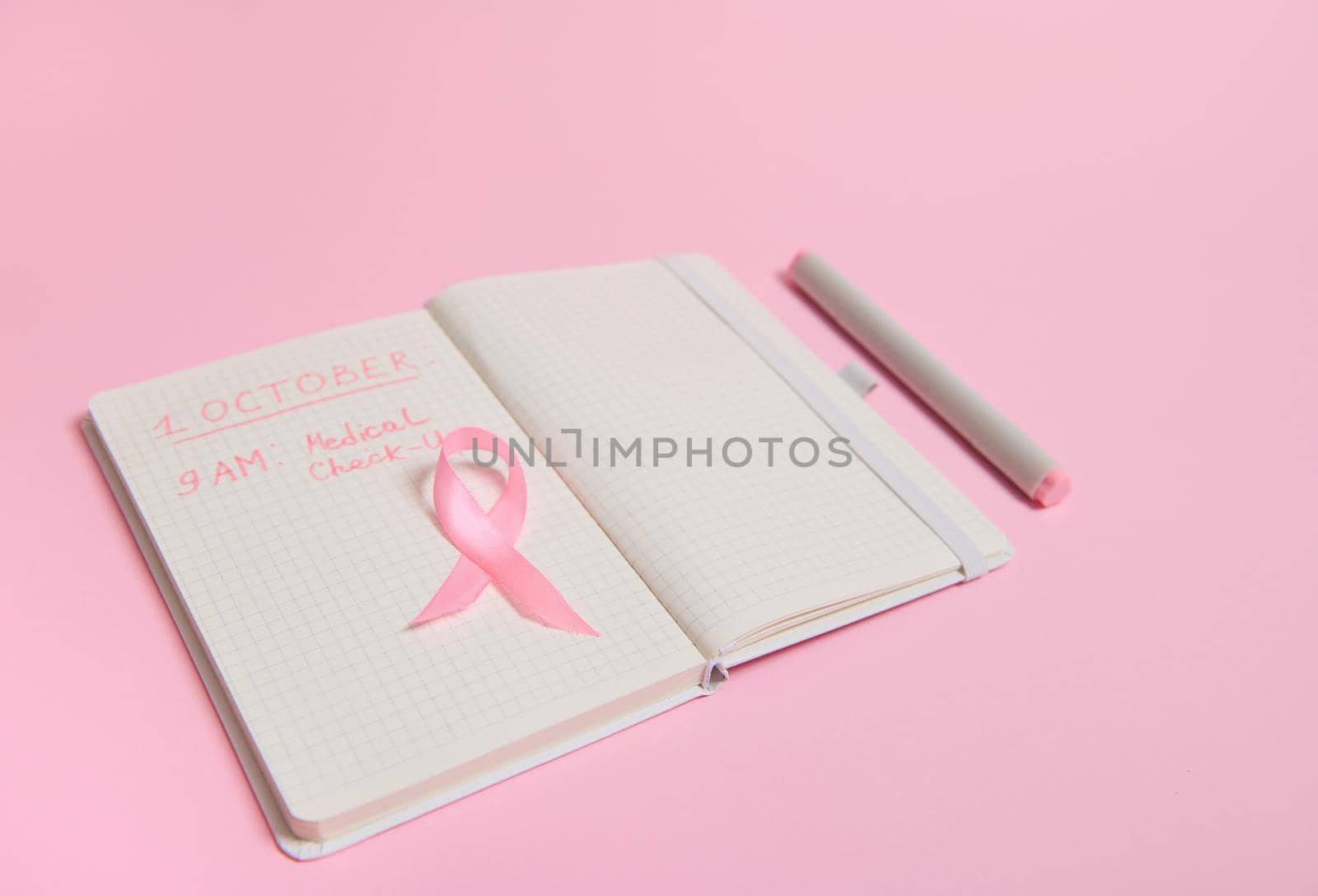 High angle view of a pink ribbon, on opened diary with inscriptions reminding of a medical check-up. October 1st, World Breast Cancer Day, Women's health care and medical concepts.