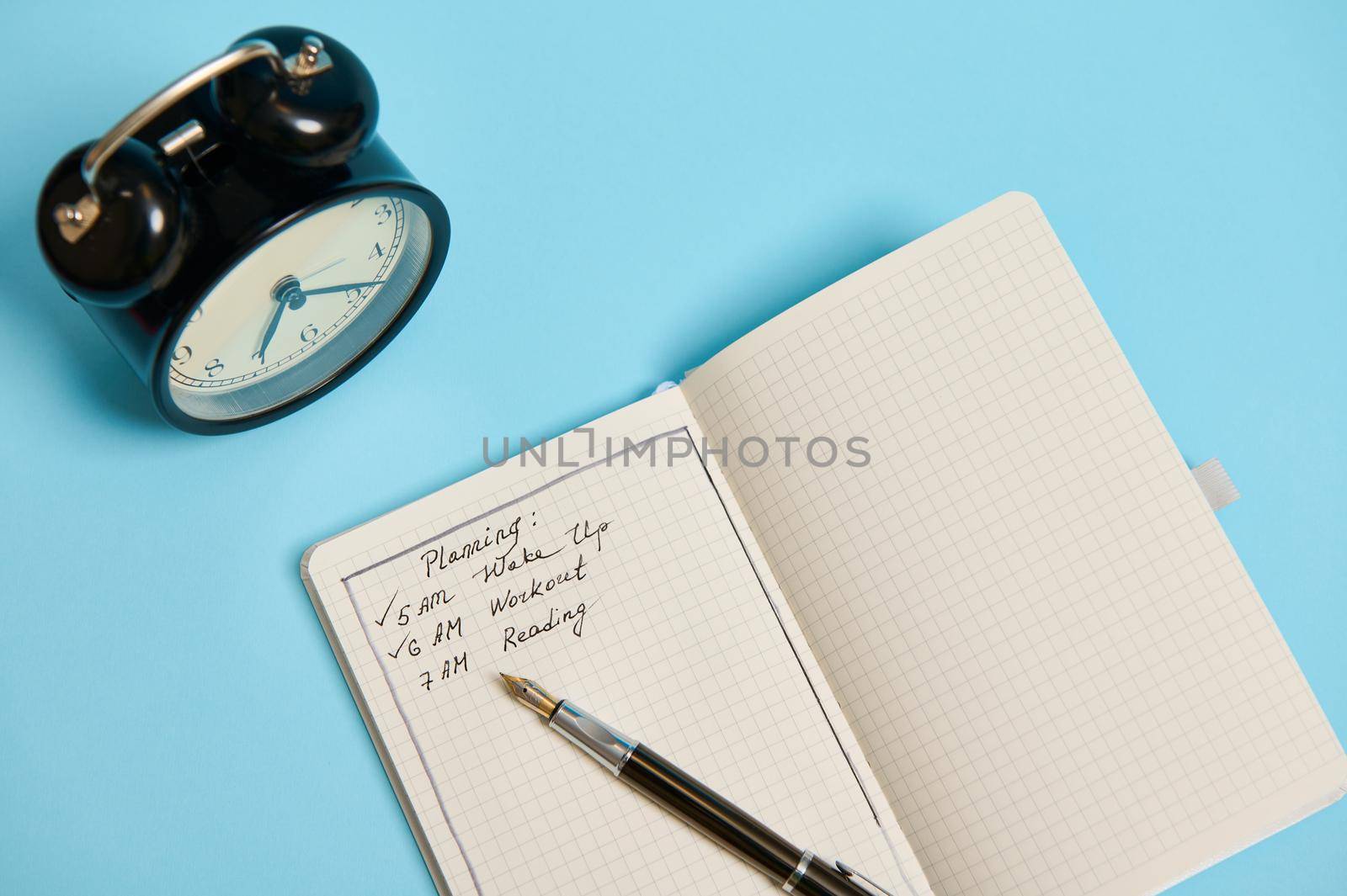 High angle view of an open organizer with plans for the day, ink pen and alarm clock on blue background with copy space