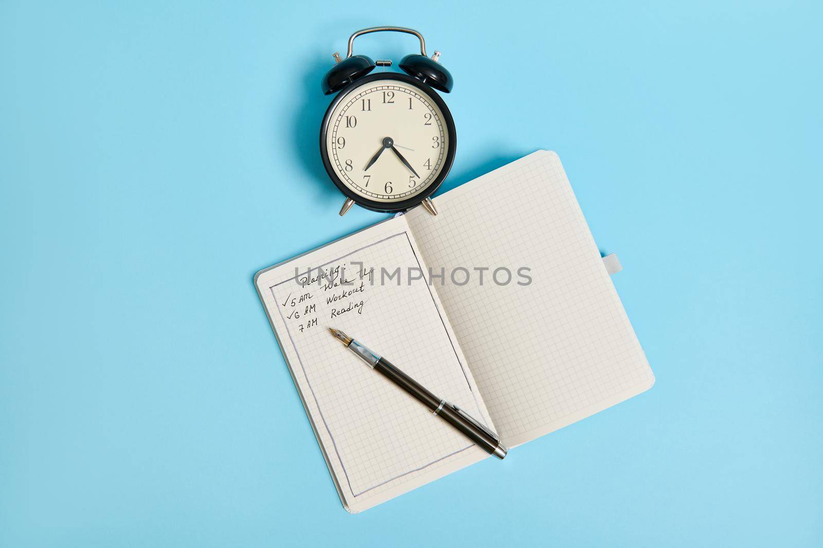 Flat lay of an open organizer with plans for the day, ink pen and alarm clock on blue background with copy space. Time management, planning and business concept