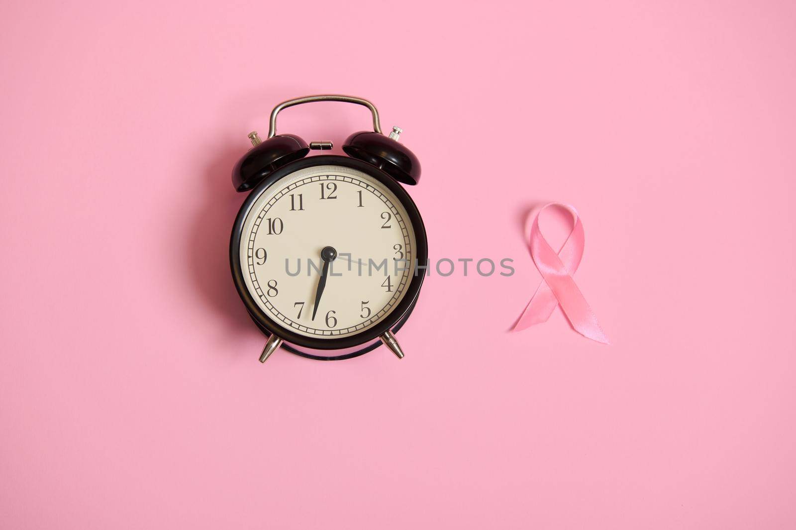 Don't miss the time, flat lay composition with an alarm clock and a pink ribbon. Breast Cancer Awareness, medical concept isolated on pink background with copy space. Women's health care concept. by artgf