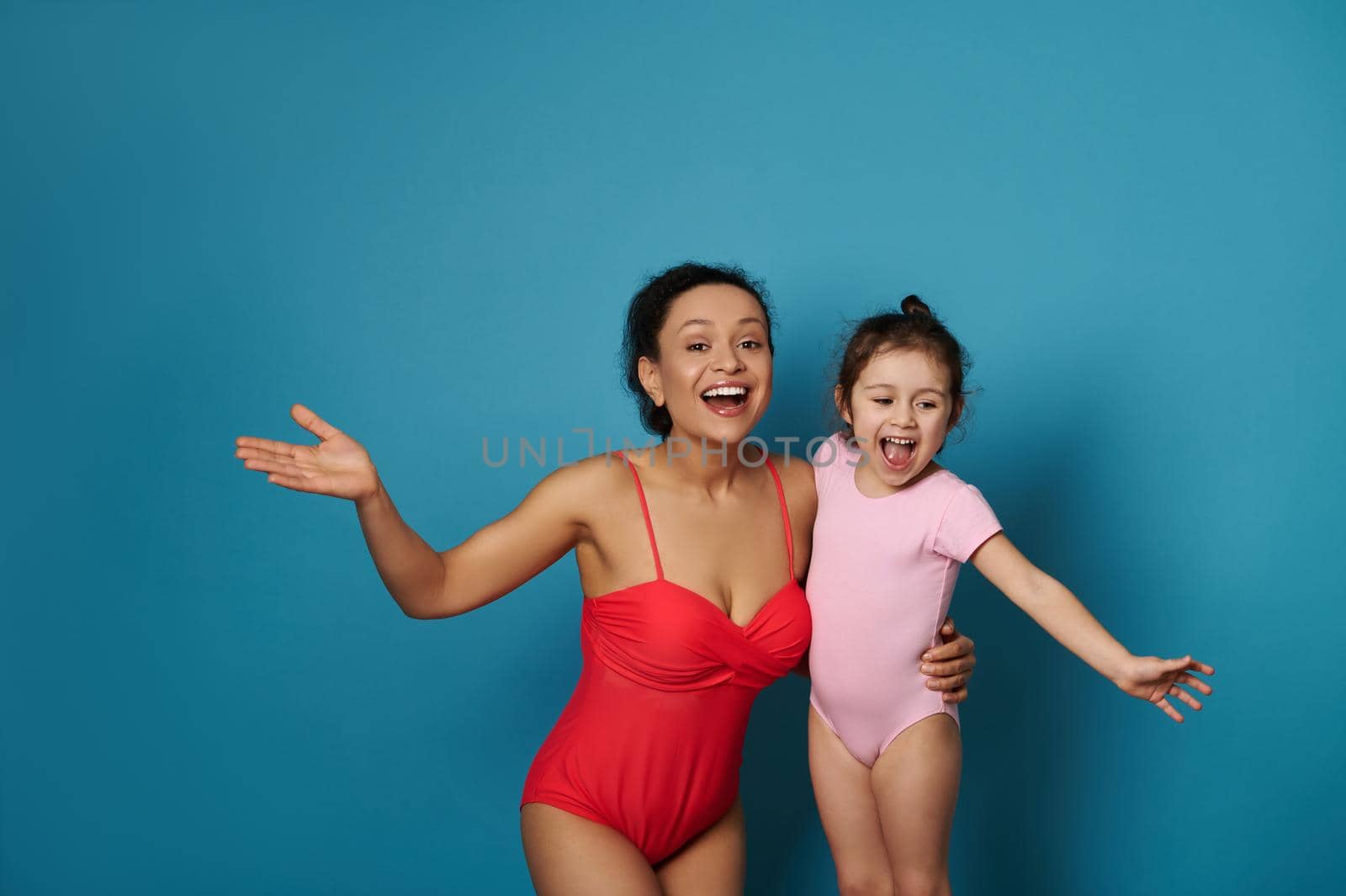 Mom and daughter in swimsuits smiling while posing over blue background with space for text