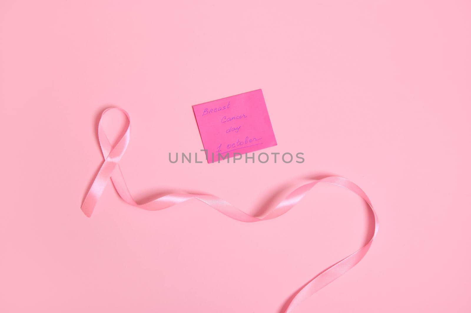Flat lay of a long pink ribbon with endless one end, and a paper note with lettering 1 October Breast Cancer Awareness Day , isolated on pink background with space for text