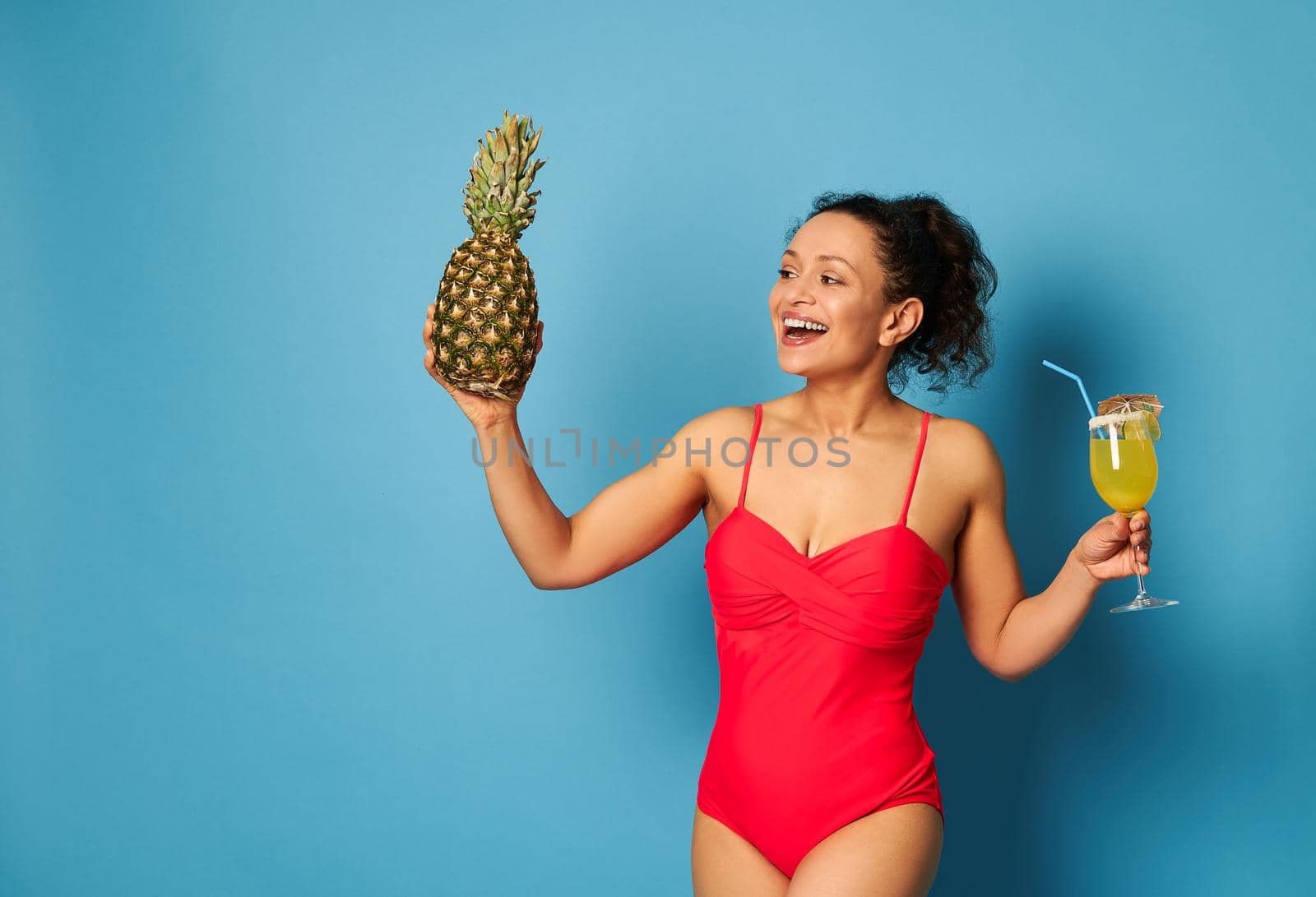 Muscular build woman in red swimsuit with a pineapple and cocktail in hands posing over blue background with copy space