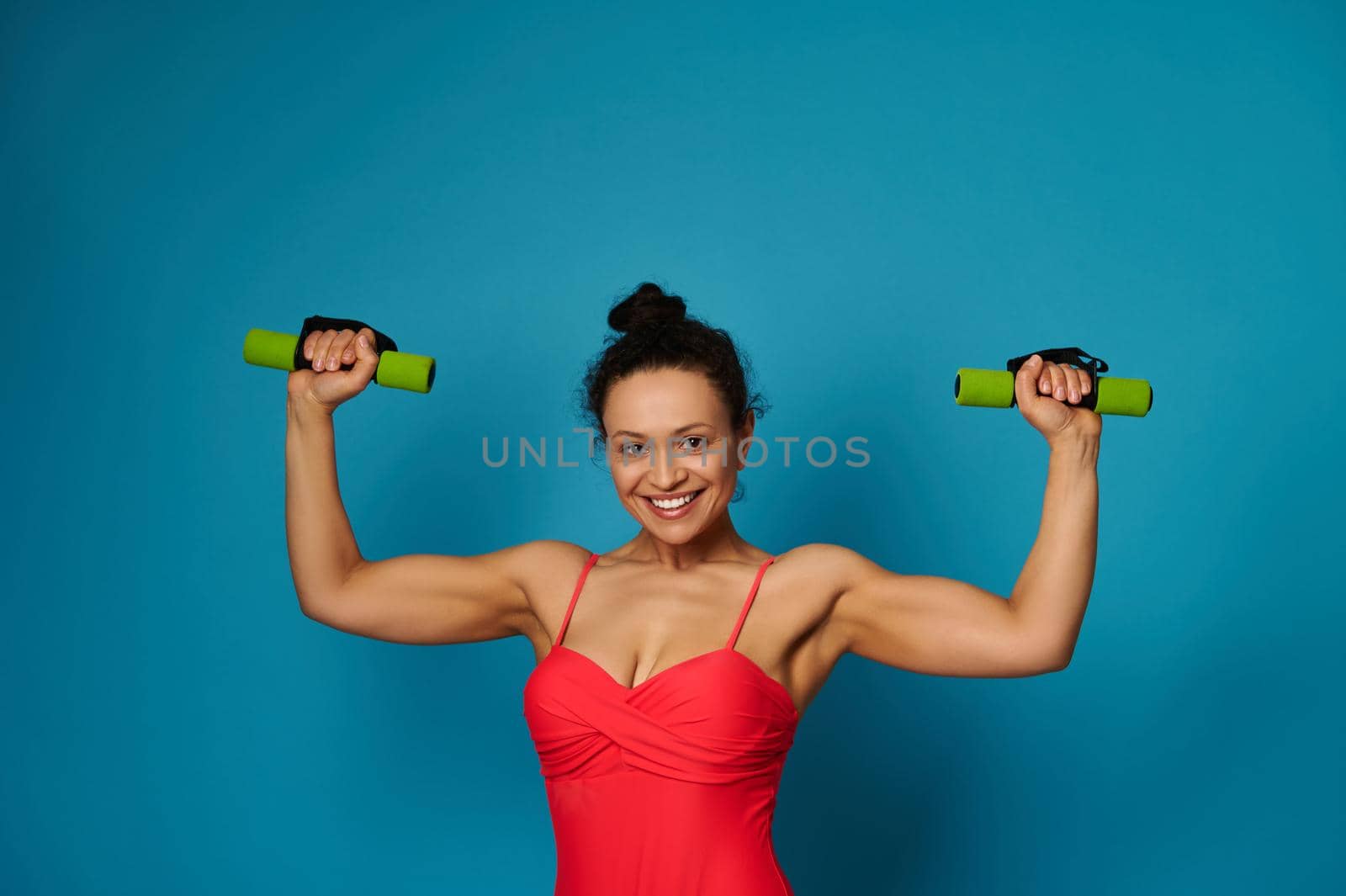 Smiling young athletic woman posing with dumbbells on blue background with copy space by artgf