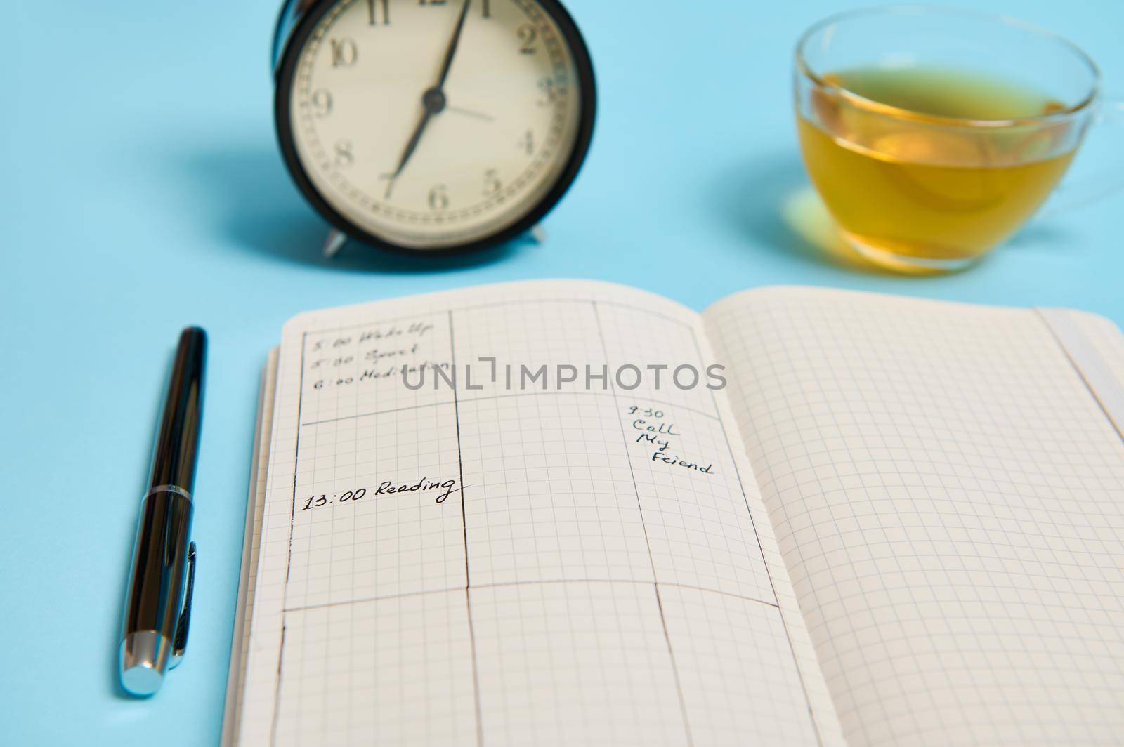 Time management, deadline and schedule concept: alarm clock on schedule plan, organizer with plans, ink pen and transparent glass cup with tea on blue background with copy space for text