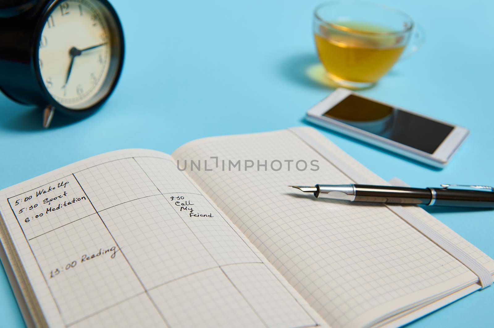 An open agenda with schedule, smartphone, ink pen, glass cup of tea and black alarm clock lie on blue surface. Color background with copy space . Time management, deadline, schedule concept by artgf