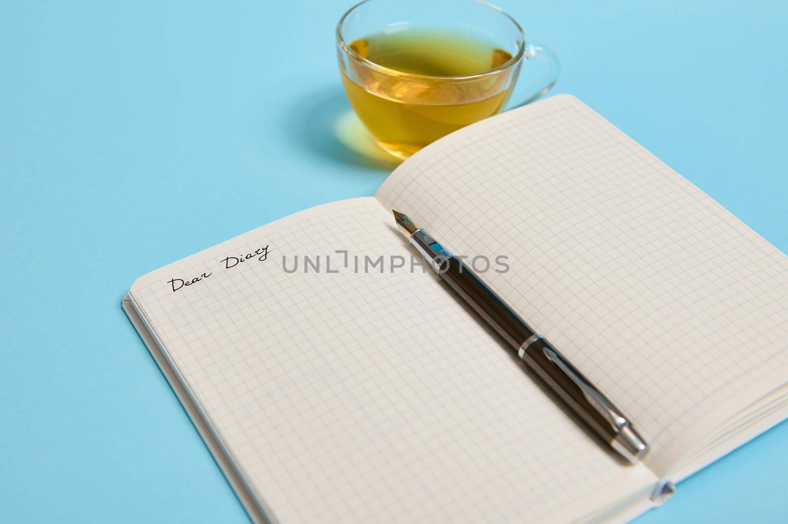 Open notepad with word Dear Diary and an ink pen next to a tea cup on blue background with copy space for text