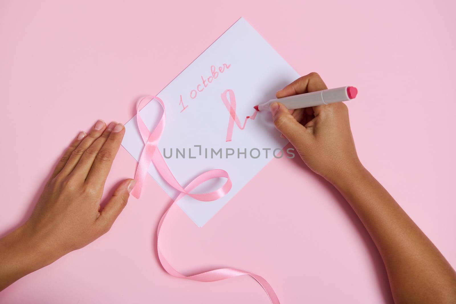 Flat lay of a woman's hand holding a felt-tip pen writes october 1 and draws on the paper a pink symbol of breast cancer awareness month, a pink ribbon with an endless end, lying on a pink background