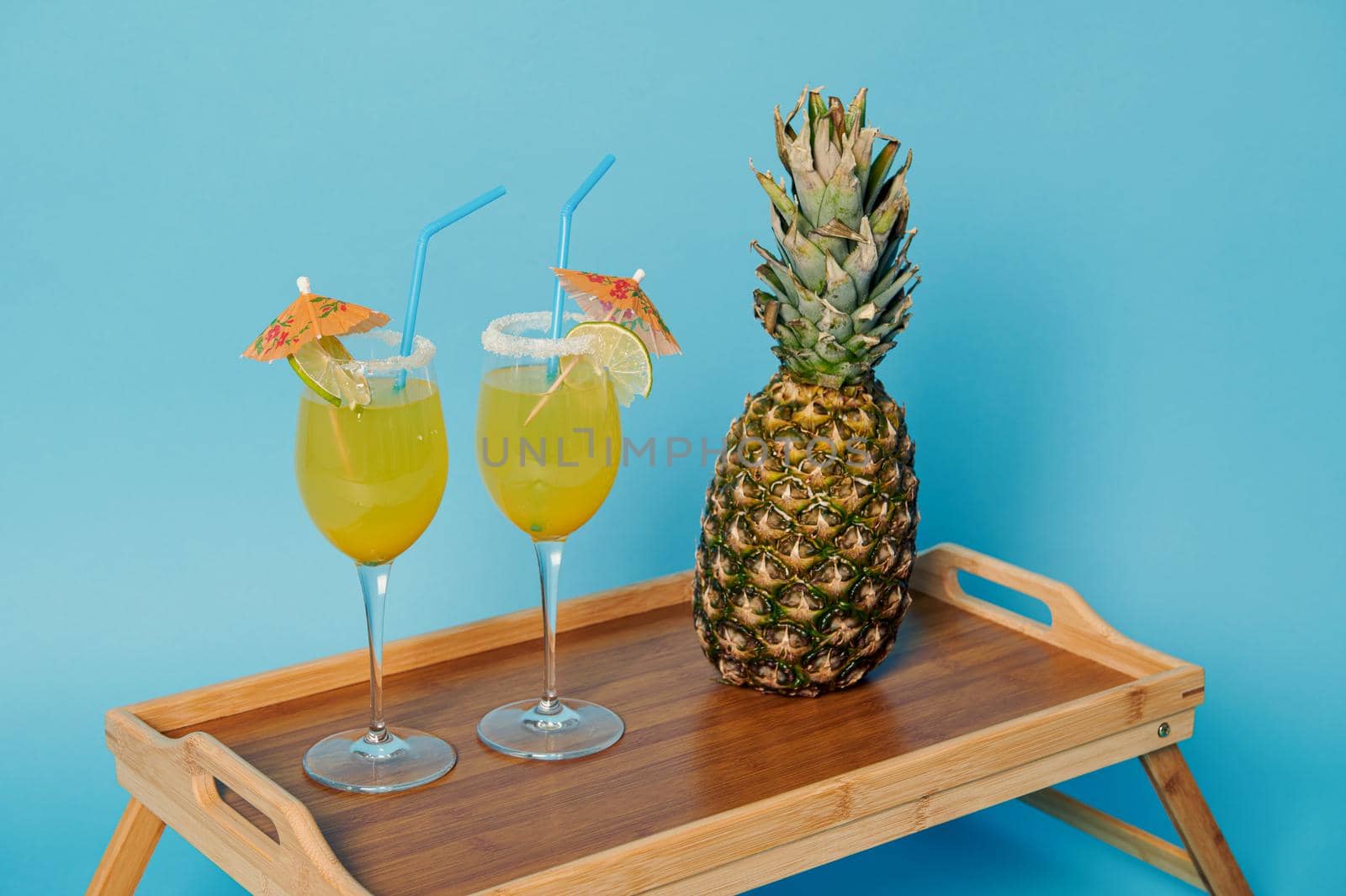 High angle view of pineapple and two glasses with exotic summer cocktails decorated with cocktail umbrella on bamboo serving tray