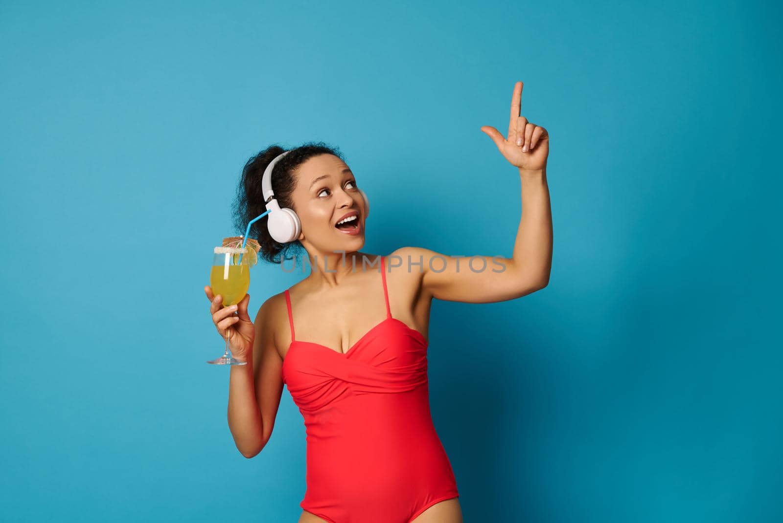 Summer concept with a cute brunette in swimsuit and headphones, holding summer cocktail and pointing finger up on blue background by artgf