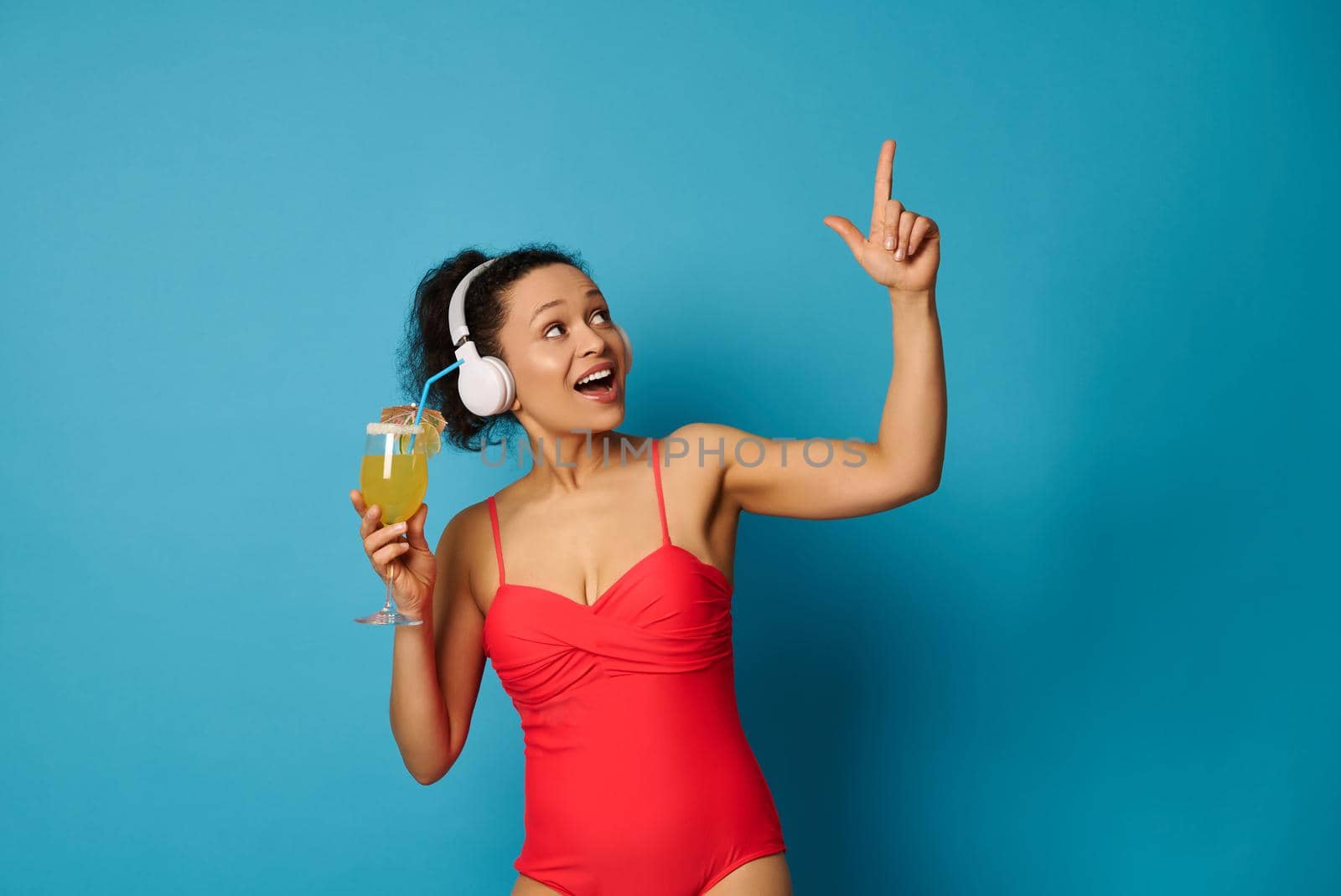 Summer concept with a cute brunette in swimsuit and headphones, holding summer cocktail and pointing finger up on blue background with copy space