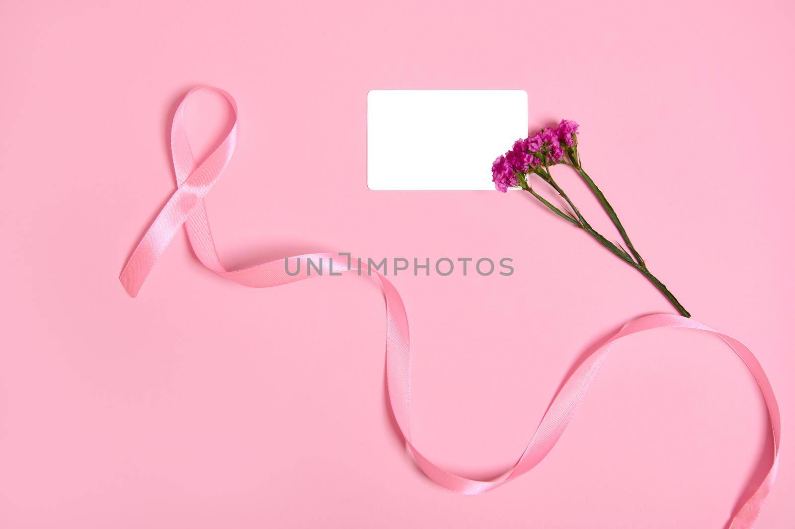 Top view of long pink ribbon with endless one end, symbol of Breast Cancer Awareness Day, a cute bouquet of flowers and an empty white blank plastic card with copy space, on pink background. Flat lay