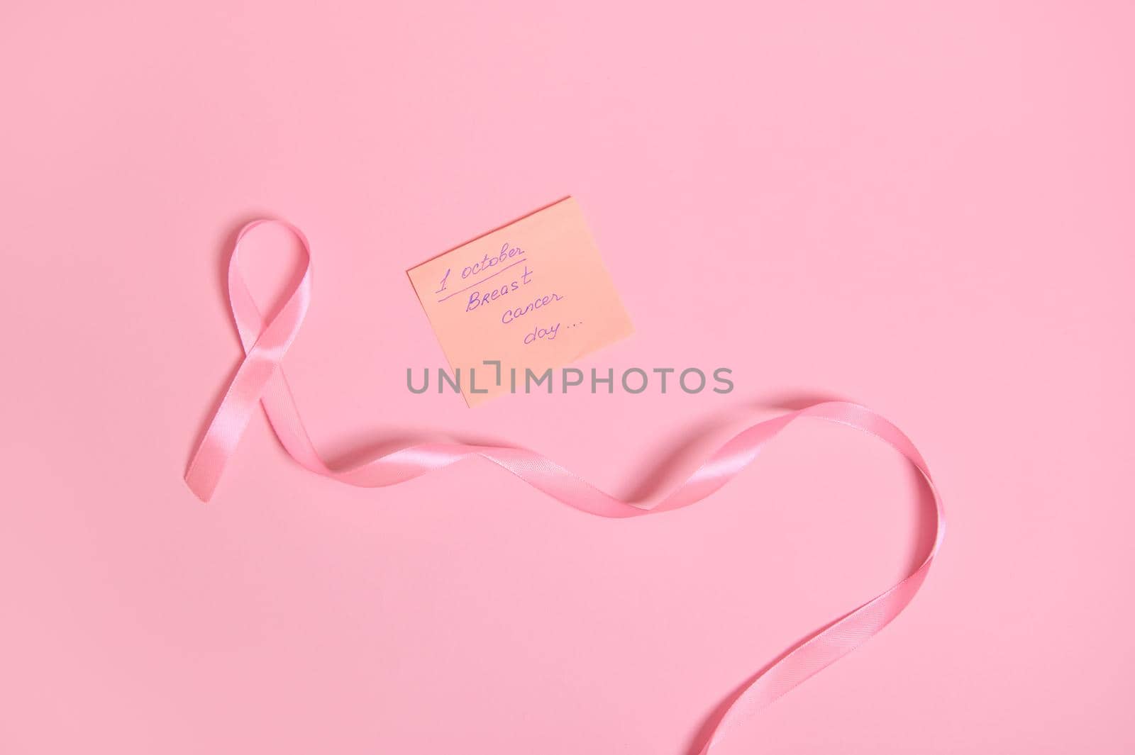 High angle view of a long pink ribbon with endless one end, and a paper note with lettering 1 October Breast Cancer Awareness , isolated on pink background with copy space. Flat lay